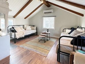 Une chambre située dans un espace de type « loft », avec deux lits en métal, une petite table ronde, une fenêtre et un appareil de climatisation.