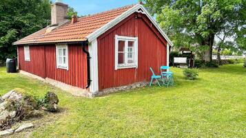 A red wooden house with a sloped roof, a chimney, and a small garden area with two blue chairs.
