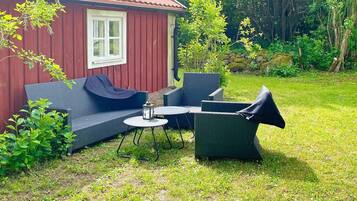 A red wooden house with a white window, a blue sofa, and a round table with chairs in a grassy backyard.