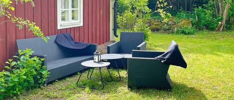 Une maison en bois rouge avec une fenêtre blanche, un canapé bleu et une table ronde entourée de chaises, le tout situé dans une cour arrière verdoyante.