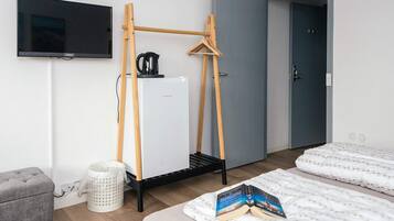 Una habitación con una cama, televisión, un refrigerador pequeño, una tetera y un perchero de madera.