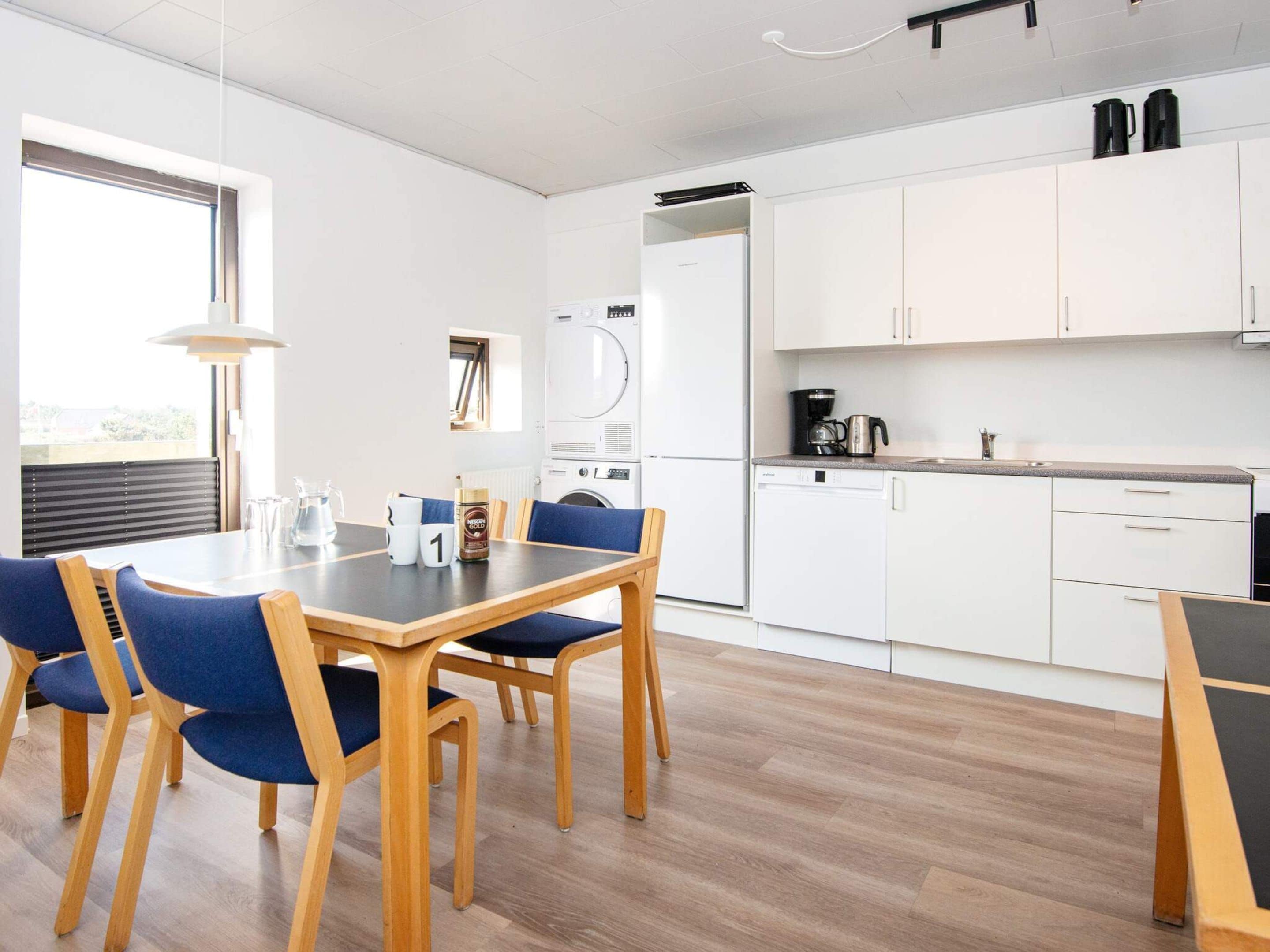 A modern kitchen with white cabinets, a dishwasher, and a coffee maker. There is a dining table with blue chairs and a window providing natural light.