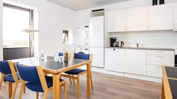 A modern kitchen with white cabinets, a dishwasher, and a coffee maker. There is a dining table with blue chairs and a window providing natural light.
