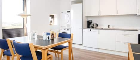 A modern kitchen with white cabinets, a dishwasher, and a coffee maker. There is a dining table with blue chairs and a window providing natural light.