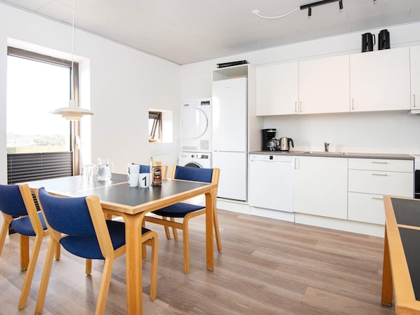 A modern kitchen with white cabinets, a dishwasher, and a coffee maker. There is a dining table with blue chairs and a window providing natural light. - 3 Person Holiday Park Home in Ulfborg (Ulfborg)