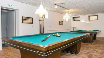 A pool table with a cue stick and balls, a second table in the background, and a ceiling with hanging lights.