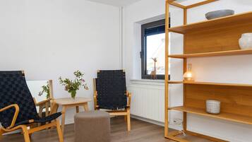 A modern living room with two black chairs, a small round ottoman, a wooden bookshelf, a small table with a vase of flowers, and a window with a view of the outside.