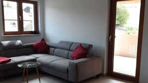 A living room with a grey sofa, two red cushions, a glass coffee table, and a sliding glass door leading to a balcony. - Holiday Apartment Wine bar (Königheim)