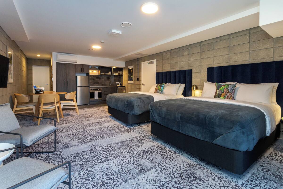 Quarto de hotel moderno com uma cama grande, sofá, área de jantar e cozinha compacta.
