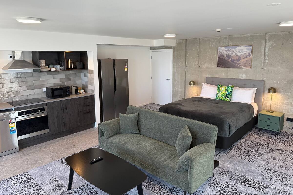Quarto de hotel moderno com cozinha compacta, cama com cabeceira, sofá e uma mesa pequena.