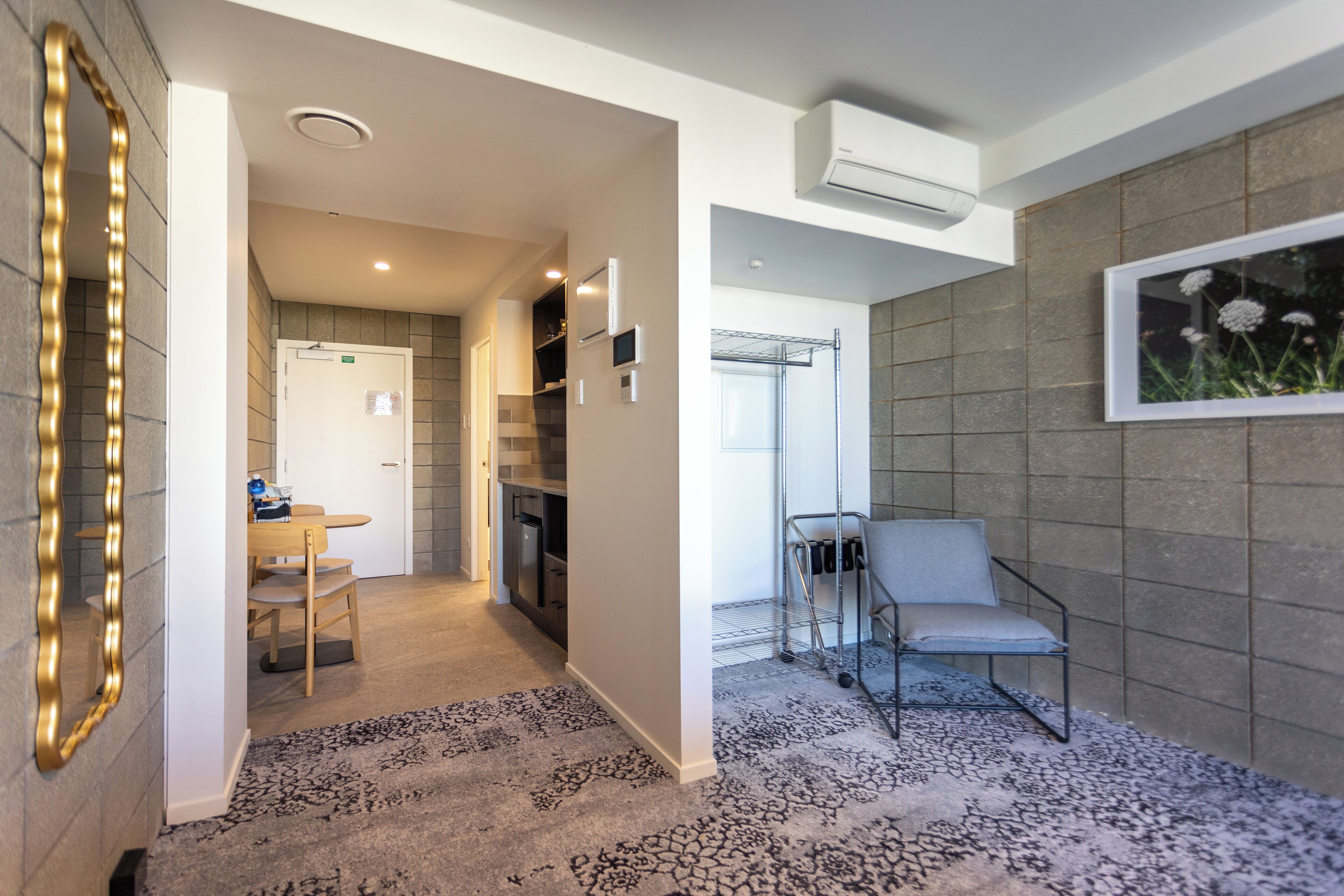 A hotel room with a modern design, featuring a large mirror, a seating area with a chair, a small table, and a kitchenette.