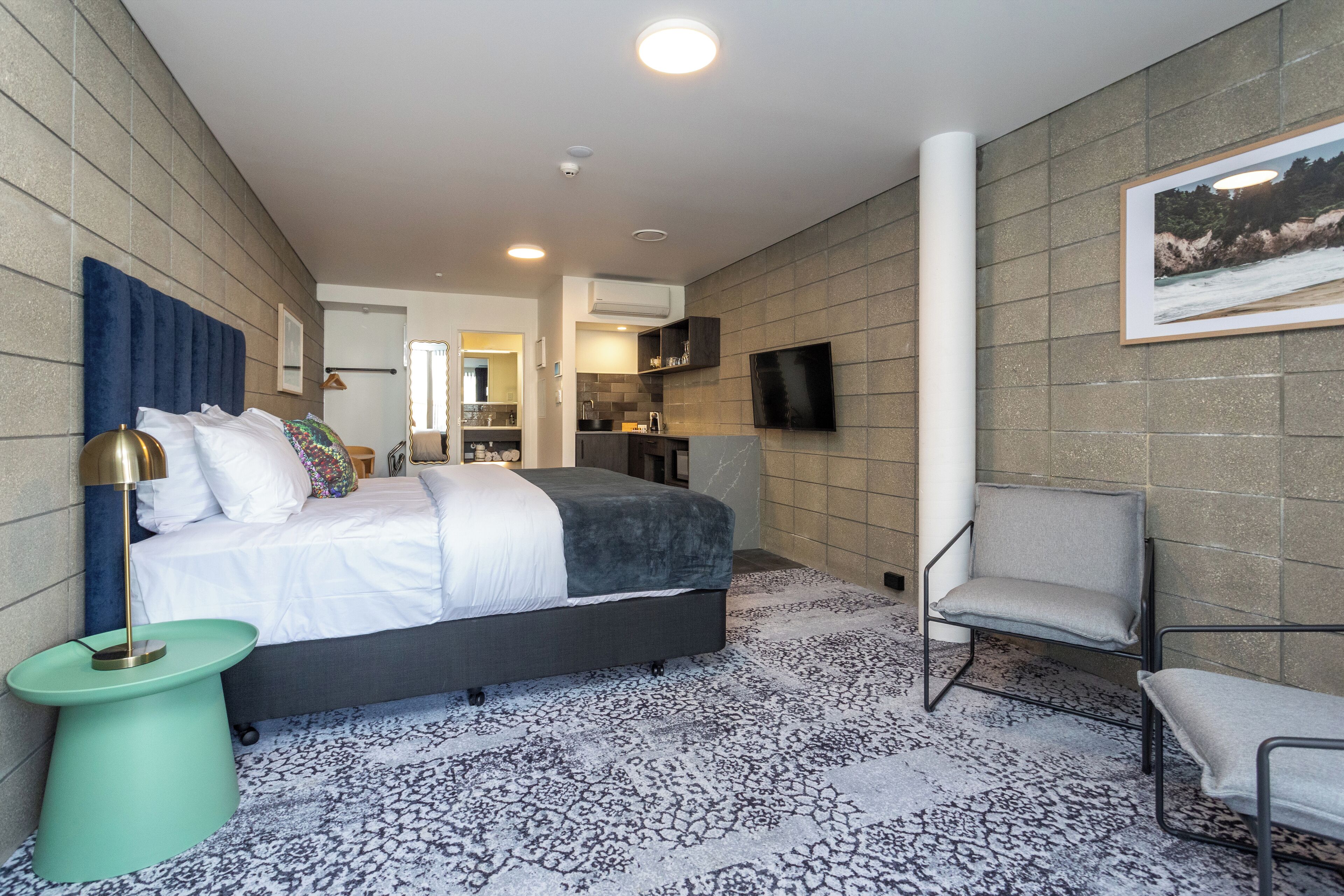 Ein modernes Hotelzimmer mit einem großen Bett, einer Sitzecke inklusive Sessel, einem kleinen Beistelltisch, einem flachbildfernseher an der Wand und einem gemusterten Teppich.