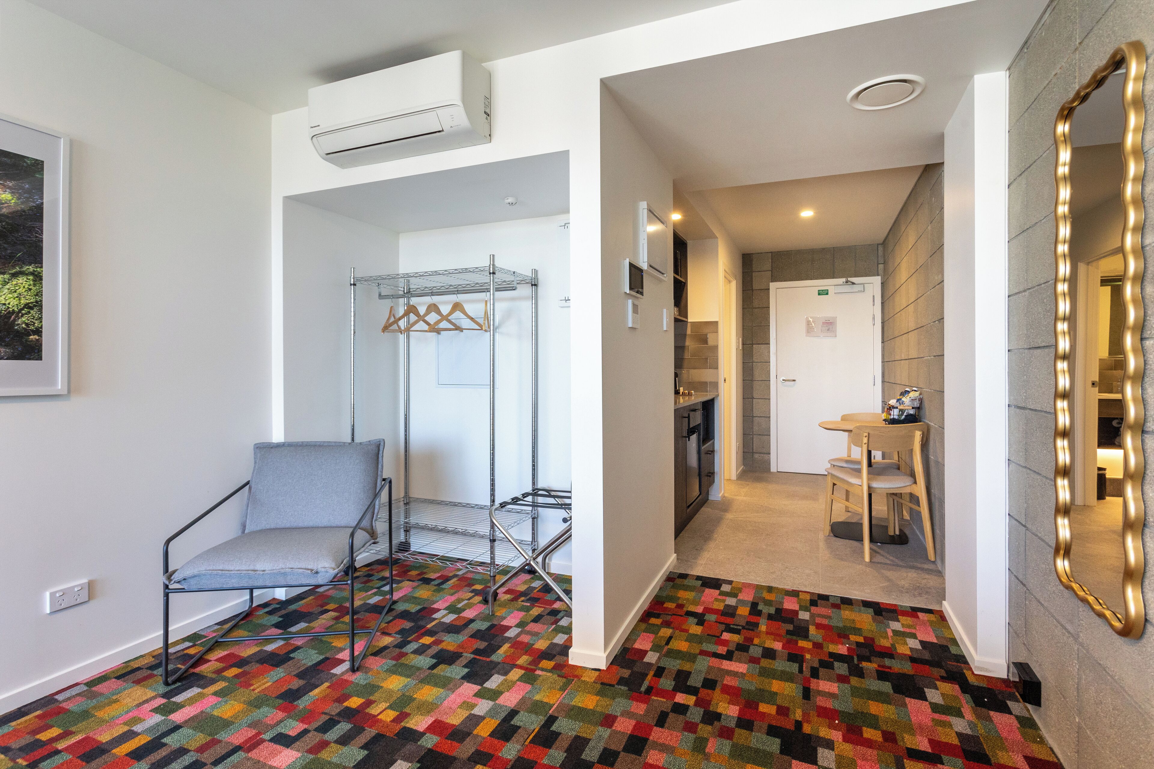 Habitación de hotel con una alfombra de diseño colorido, una silla gris, un tendedero metálico, una puerta blanca que da a un baño y un espejo con marco dorado.