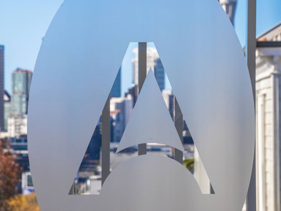 A large, white, circular sign with a triangular cutout, set against a cityscape with buildings and a tower in the background.