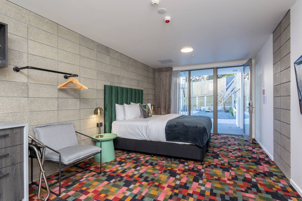 Quarto de hotel moderno com uma cama grande, uma cadeira, uma pequena mesa lateral e um tapete colorido.