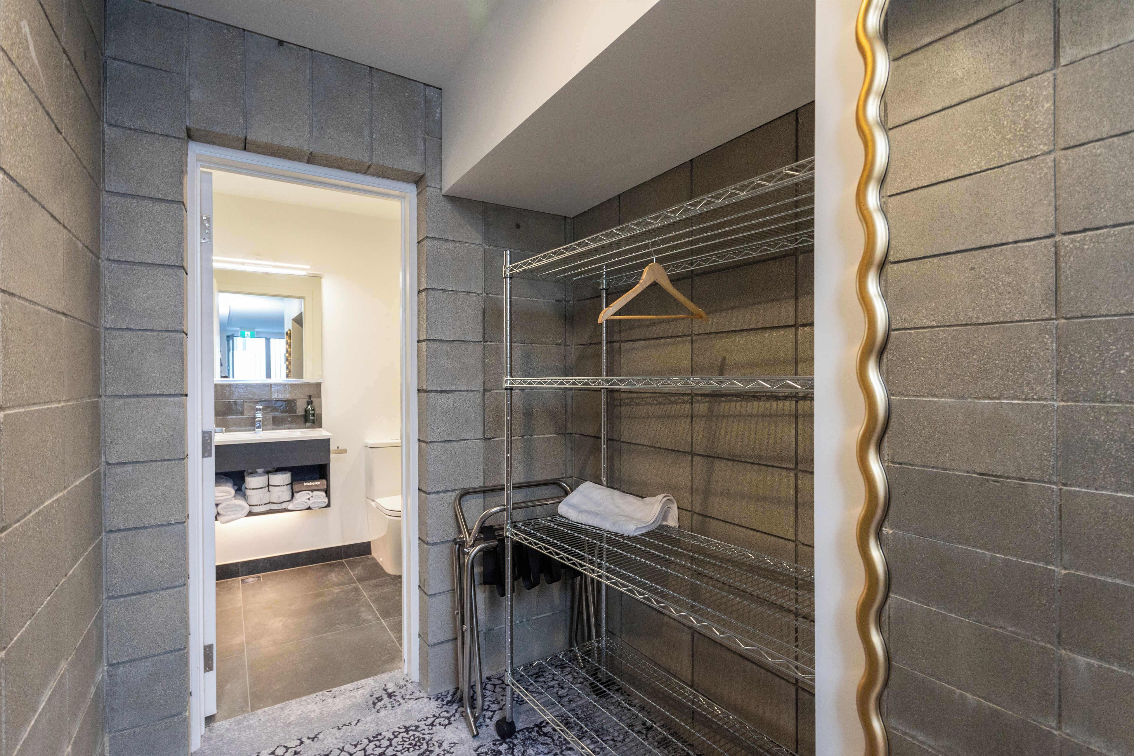A modern bathroom with grey tiled walls, a white door leading to a bathroom area, and metal shelves with a towel rack.
