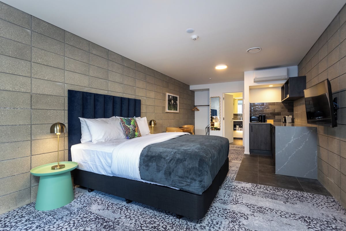 Quarto de hotel moderno com uma cama grande, uma mesinha de apoio, cozinha compacta e uma televisão de tela plana fixada na parede.