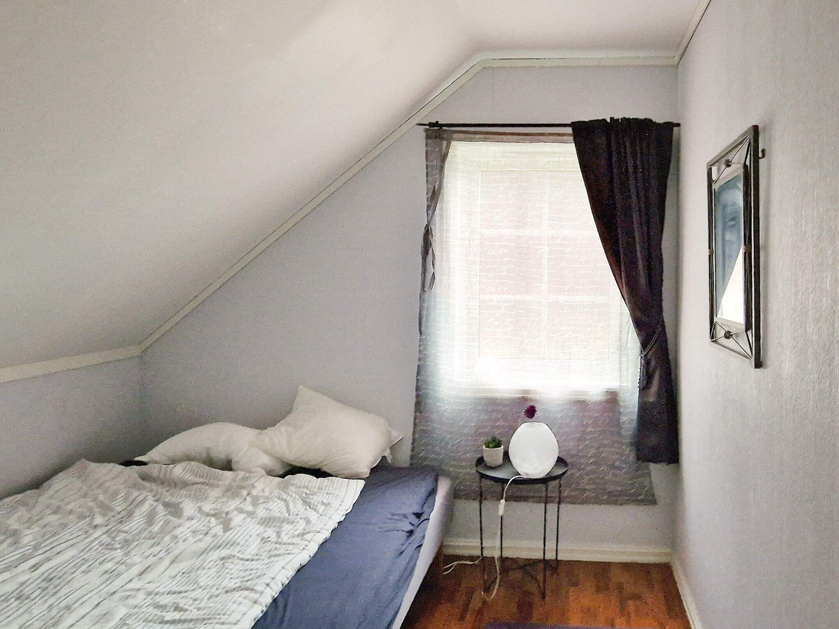 Ein kleines, gemütliches Schlafzimmer mit schrägen Wänden, einem Einzelbett mit Kissen, einem Nachttisch mit Lampe und einem Fenster mit Vorhängen.