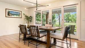 Uma área de refeições com mesa e cadeiras de madeira, luminária pendente e uma grande janela com vista para o verde.