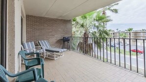A balcony with two lounge chairs, a small table, and a view of palm trees and a street. - Padre Oasis #302 (South Padre Island)