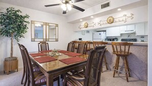 A dining area with a wooden table, chairs, a ceiling fan, a potted plant, and a kitchen in the background. - Padre Oasis #302 (South Padre Island)