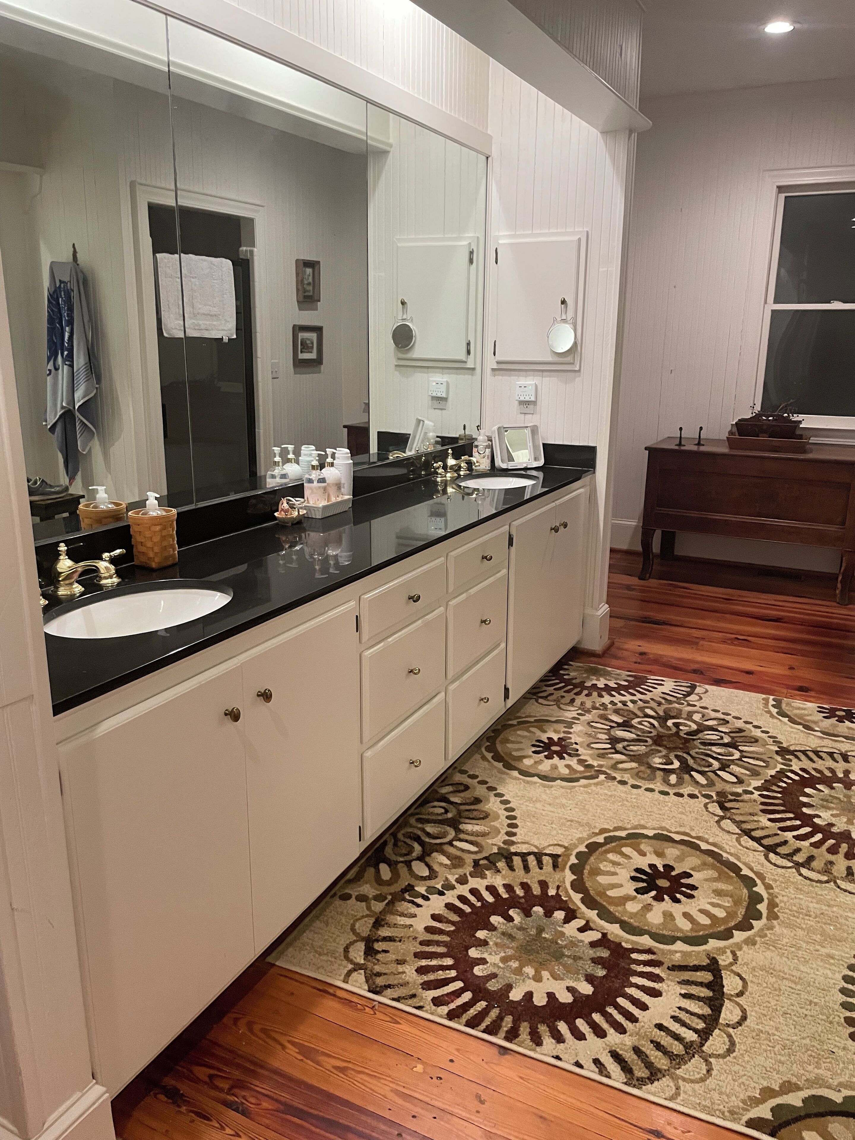 A bathroom with a large mirror, a black countertop, a white sink, and a wooden floor.
