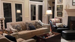 Een woonkamer met een beige bank, een donkerbruine fauteuil, een houten salontafel en een kamerkleed met dessin. Er is een plafondventilator, een open haard en lijstwerken aan de muur.