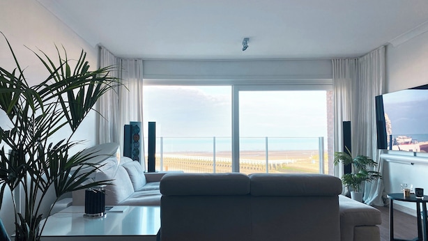 A modern living room with a large window, a sofa, a coffee table, and a TV. - Apartment in Nieuwpoort Near the Beach (Nieuwpoort)