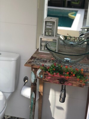 Kylpyhuone, jossa on maalaishenkinen puinen pesuallas, lasinen kulho ja wc-istuin.