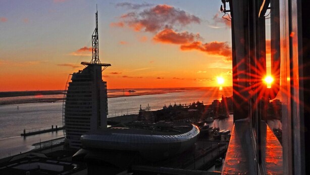 A sunset view from a high-rise building overlooking a coastal area with a distinctive modern building and a bridge.
