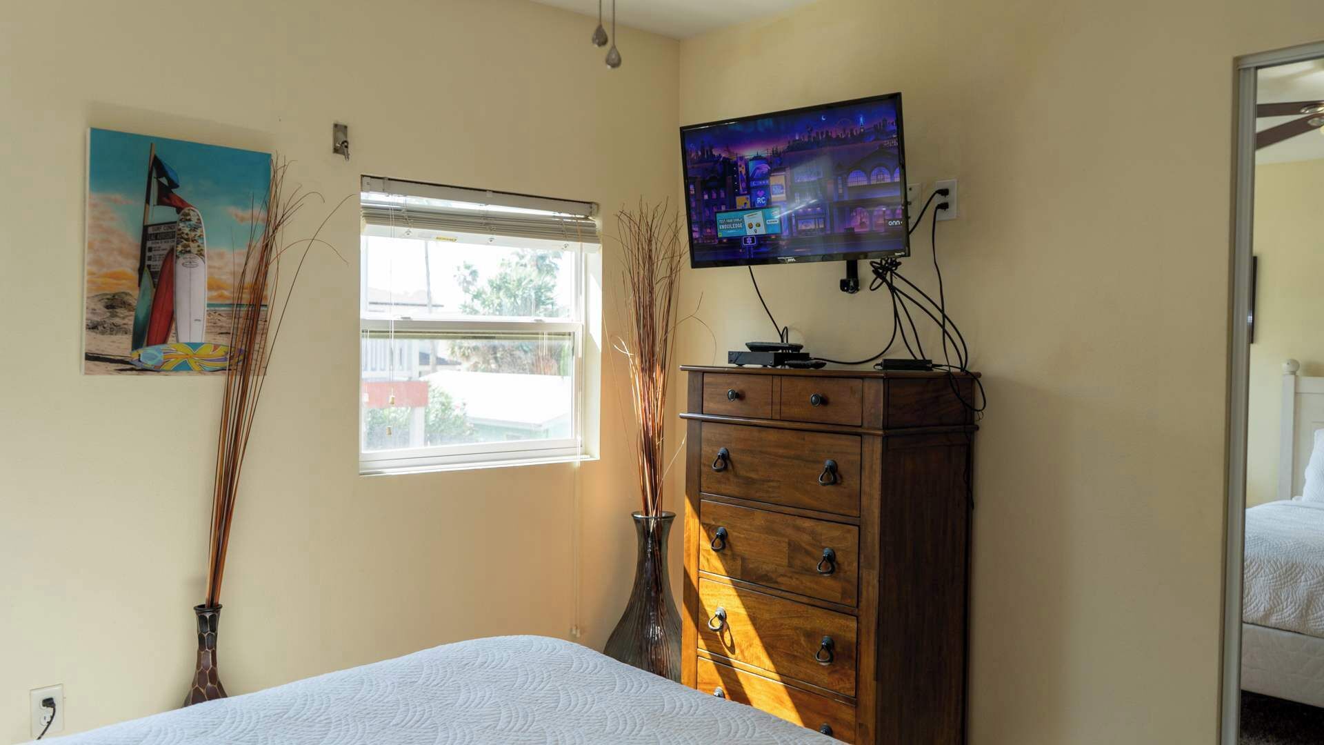 Un dormitorio con cama, cómoda, televisor montado en la pared, ventana y un cuadro.