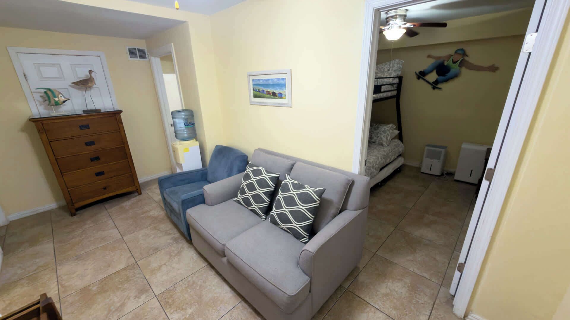 A living room with a grey sofa, a wooden dresser, a water dispenser, and a bunk bed in the background.