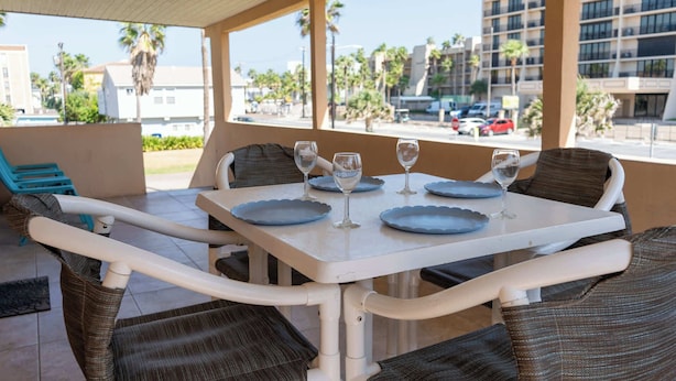 A table set for four with blue plates and wine glasses, surrounded by wicker chairs, under a covered patio with a view of palm trees and buildings. - 4 Bedroom 2 Level Home - 50 Yards From Beach (South Padre Island)