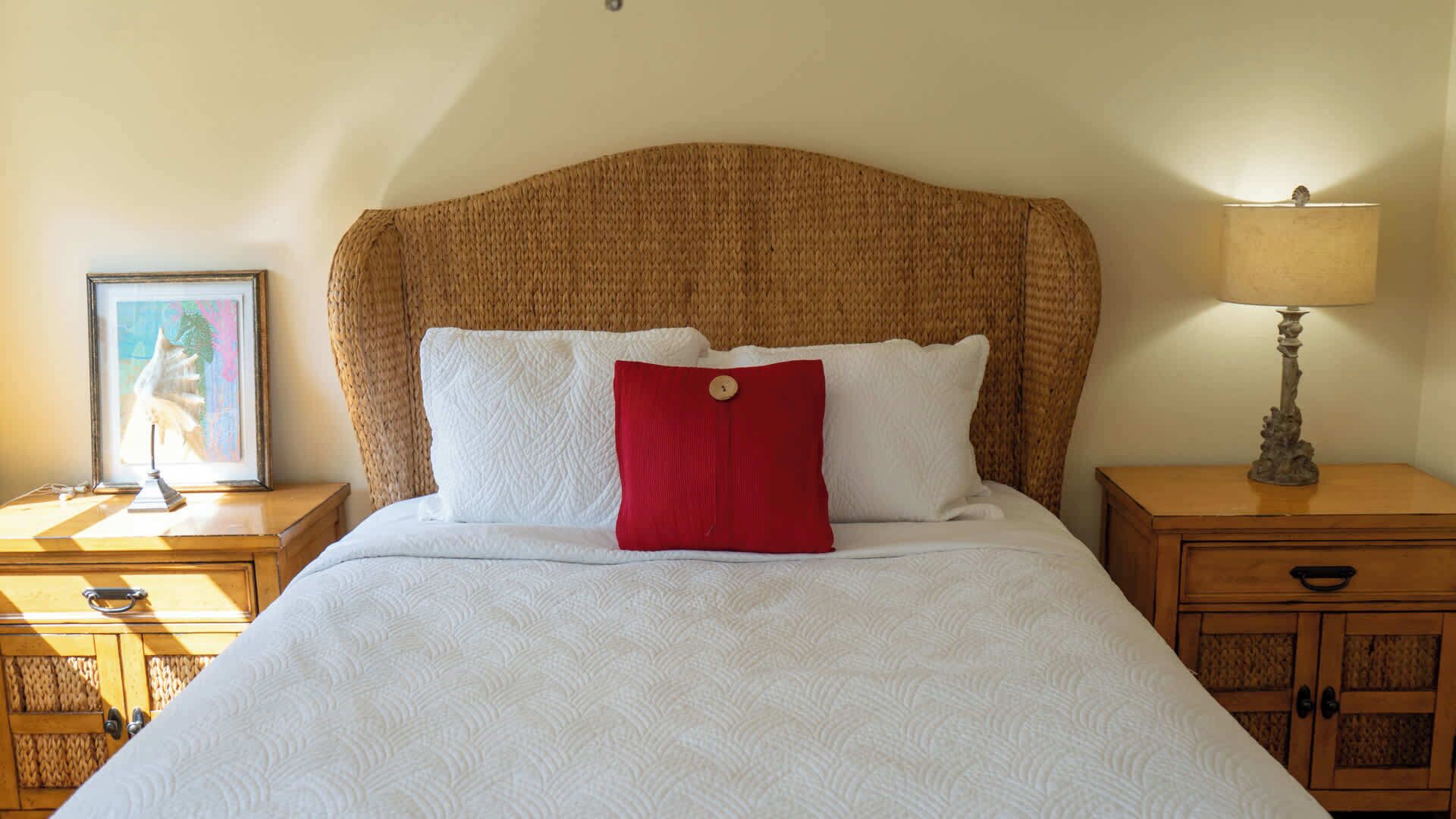 A bed with a woven headboard, white bedding, and a red pillow. There is a nightstand with a lamp and a framed picture on the left. A wooden nightstand with a drawer is on the right.