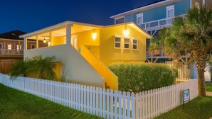A two-story house with a white picket fence, a staircase leading to the second floor, and a well-lit front porch. - 4 Bedroom 2 Level Home - 50 Yards From Beach (South Padre Island)