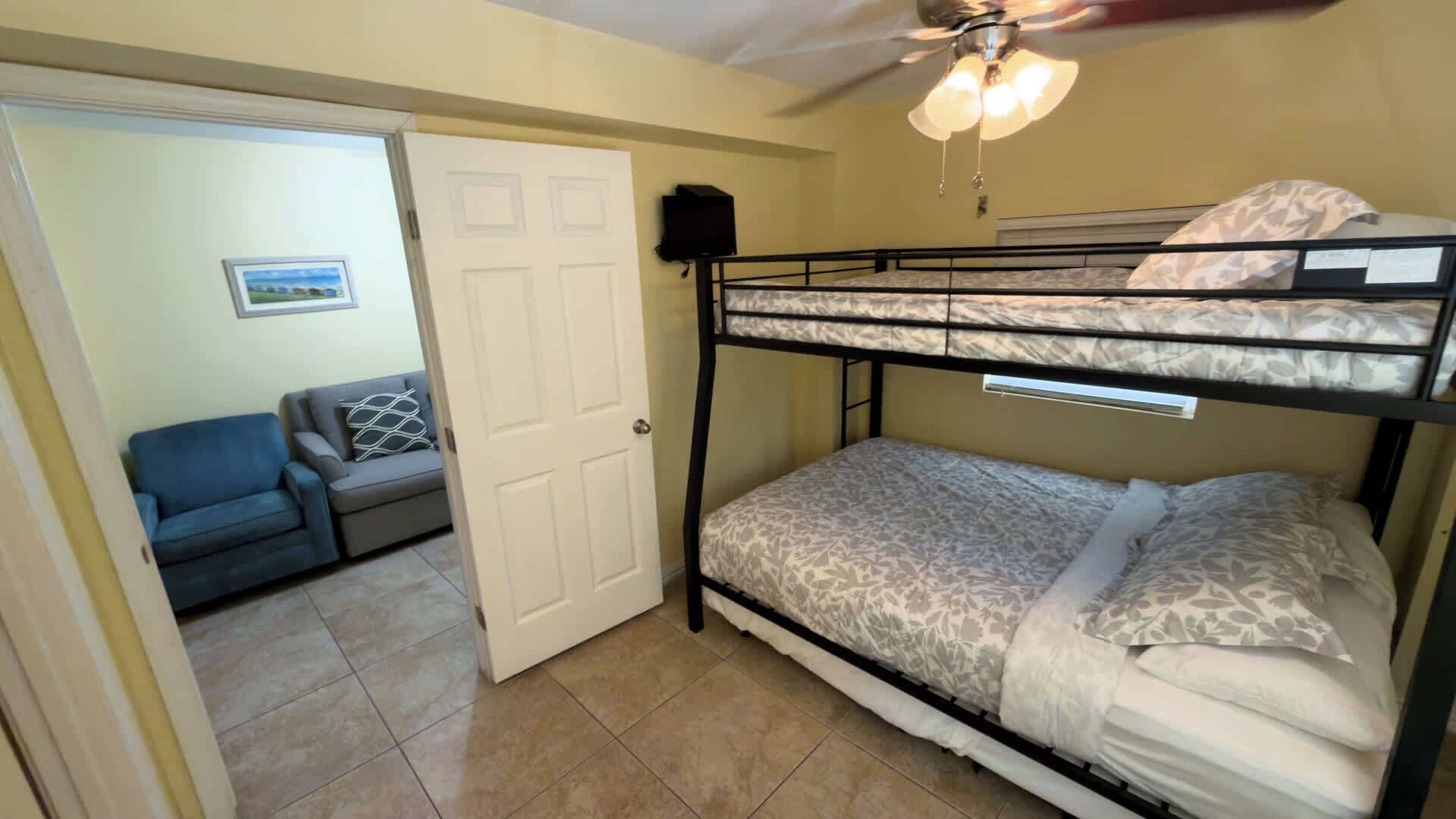 A room with a bunk bed, a door leading to another room, a ceiling fan, and a small table with a lamp.