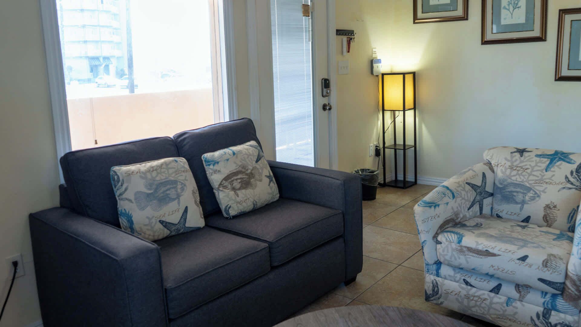 A dark grey sofa with two matching cushions, a patterned armchair, a standing lamp, and a view of the outside through a glass door.