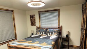 A wooden bed with a quilt, two bedside tables with lamps, a window with blinds, and a ceiling light fixture. - Come stay at the quite and Cozy 
Rustic Hideaway  (Edwards)