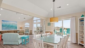 Een ruime woonkamer met een eettafel, witte stoelen, een gestreepte bank en uitzicht op de oceaan door grote ramen.