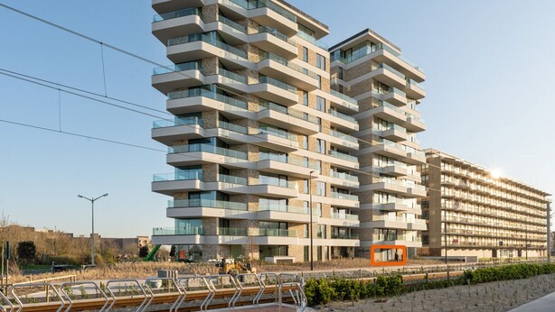Um edifício de apartamentos moderno em vários andares, com varandas, uma linha de elétrico e uma obra em construção nas proximidades.