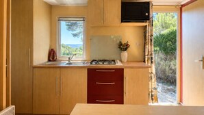 A compact kitchen with wooden cabinets, a gas stove, a microwave, and a window providing a view of greenery. - Le Romarin, mobile home with sea view (Hyères)