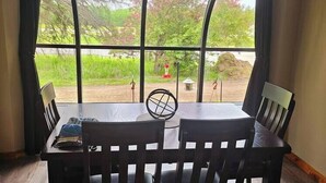 Un espace repas comprenant une table et des chaises, de grandes fenêtres donnant sur un paysage verdoyant et une statue, ainsi qu'un sol carrelé.