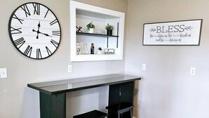 Une cuisine moderne avec un bureau en bois foncé, deux tabourets noirs, une grande horloge murale, une étagère blanche ornée d’objets décoratifs et un panneau encadré sur lequel est écrit « Bénédictions ».