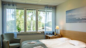 A hotel room with a bed, a chair, a nightstand with a lamp, and a window with a view of trees and a building. - Lungsunds hotell & camping (Storfors)