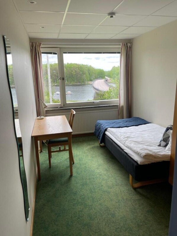 Ein kleines Hotelzimmer mit einem Bett, einem Schreibtisch und einem Stuhl. Ein Fenster bietet Blick auf ein Gewässer und Grünflächen.