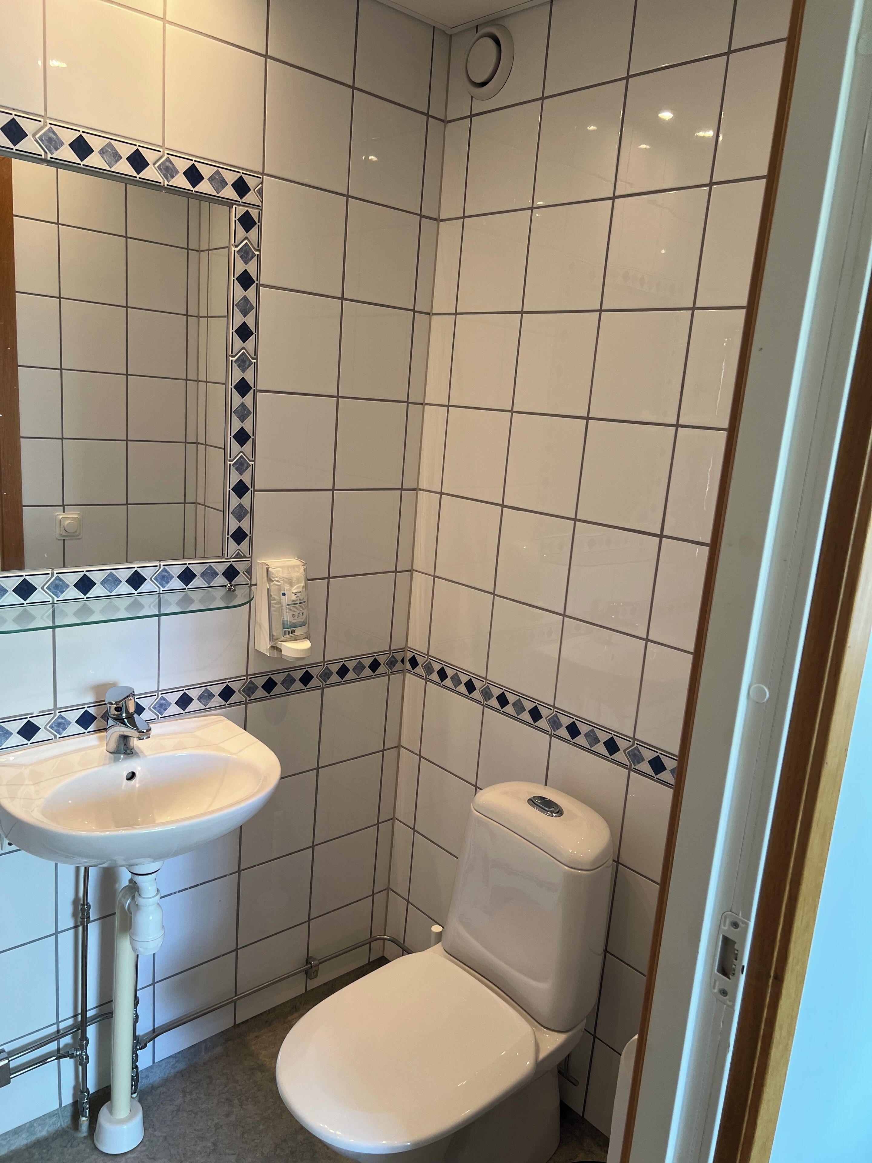 Une petite salle de bain avec un lavabo, des toilettes et un miroir, le tout de couleur blanche. Les murs sont recouverts de carreaux aux motifs blancs et bleus.