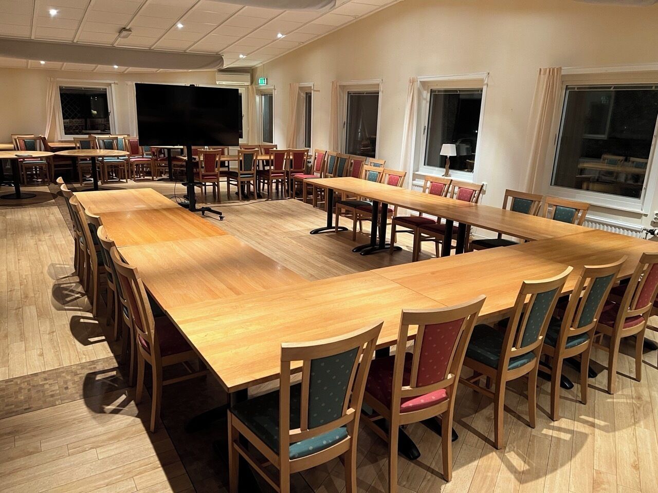 Een vergaderzaal met een lange houten tafel en meerdere stoelen, voorzien van een flatscreen televisie en ramen.