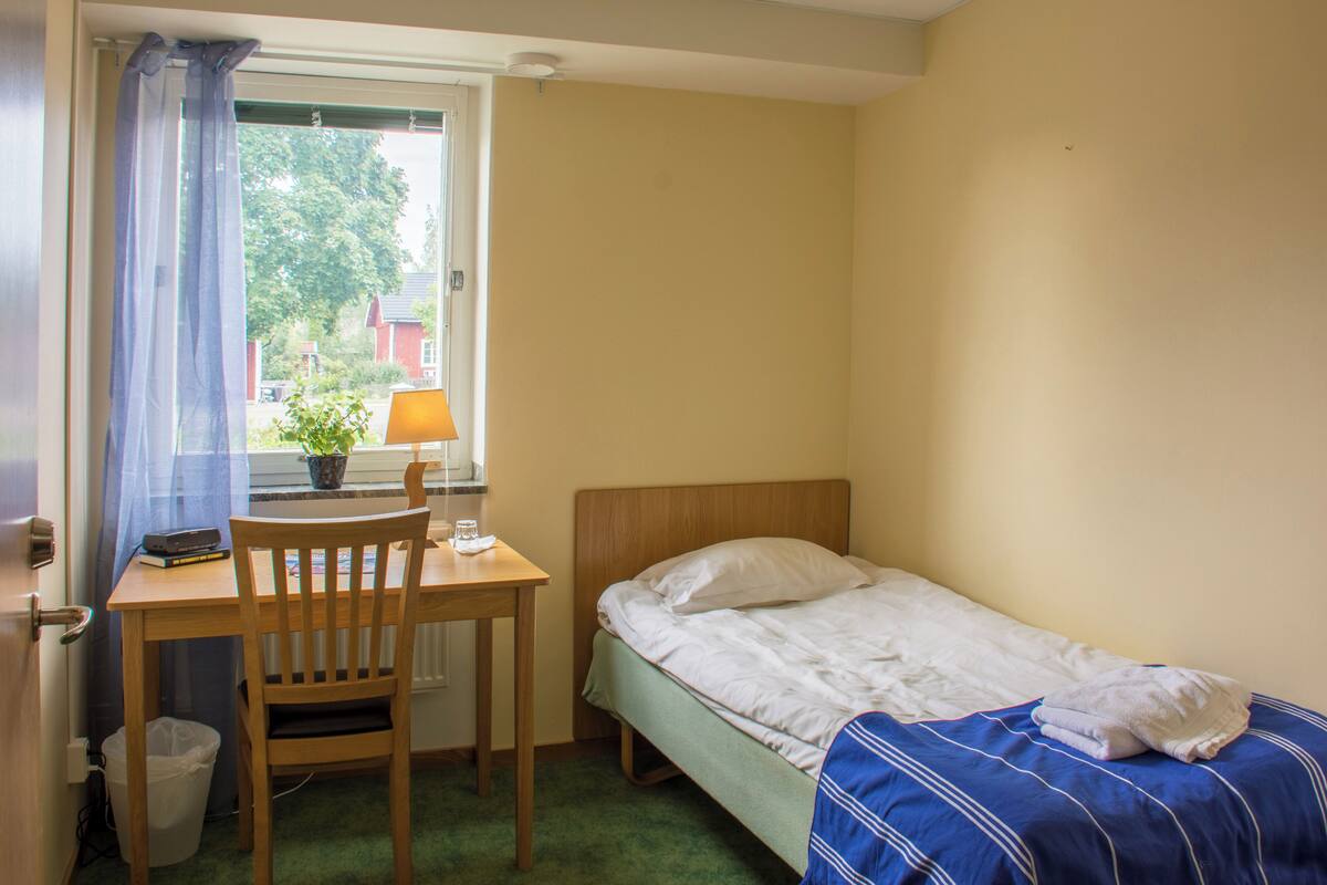 Una camera da letto modesta con un letto singolo, una scrivania in legno, una sedia, una lampada e una finestra vista casa e alberi.