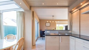 A modern kitchen with light wood cabinets, a black countertop, and a dining area with wicker chairs and a glass table. - Villa in Koksijde Near the Sea (Koksijde)