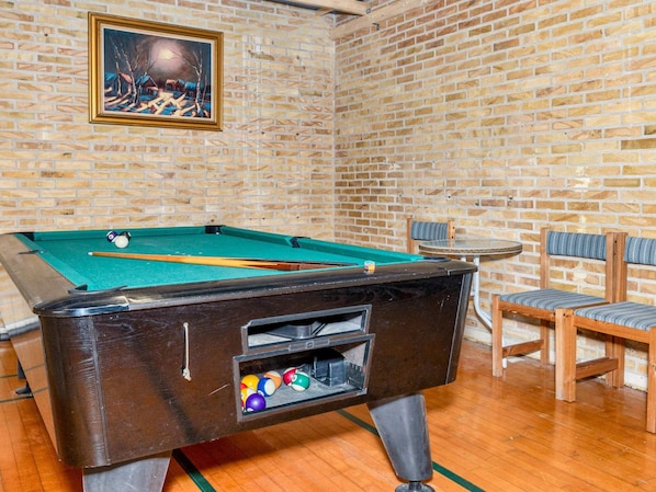 A pool table with balls and a cue stick, a framed painting on the wall, and a small round table with chairs.